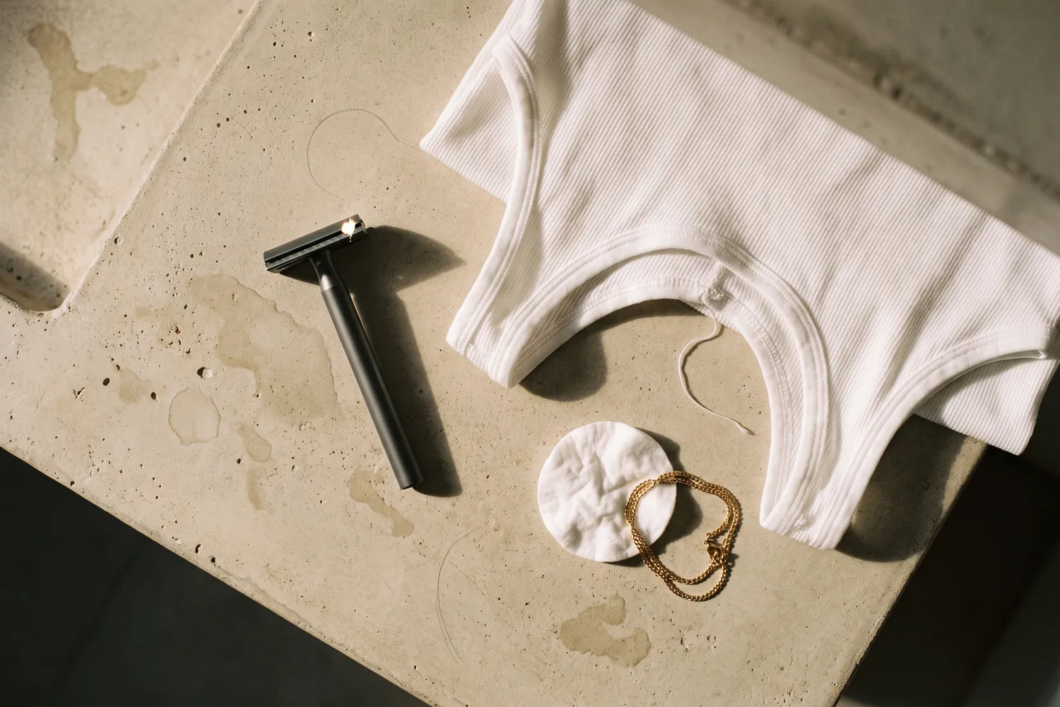 A razor, a cotton pad, a hair tie and a white tank top on a bathroom counter: the daily sources of underarm friction